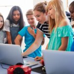 A diverse group of elementary students gathers around a table with laptops, guided by a teacher. The students are engaged and focused, collaborating on a digital project—illustrating inclusive, technology-supported learning in the classroom.