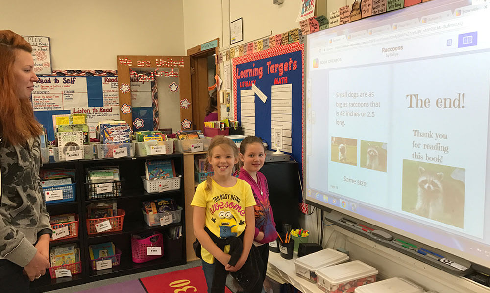 Two smiling young students stand in front of a projector screen in their classroom, presenting a digital book they created using the Book Creator app. The screen displays the final page of the book, which includes pictures of raccoons and a 'Thank you for reading!' message. A teacher watches proudly from the side, while the classroom is filled with colorful posters, books, and educational materials.